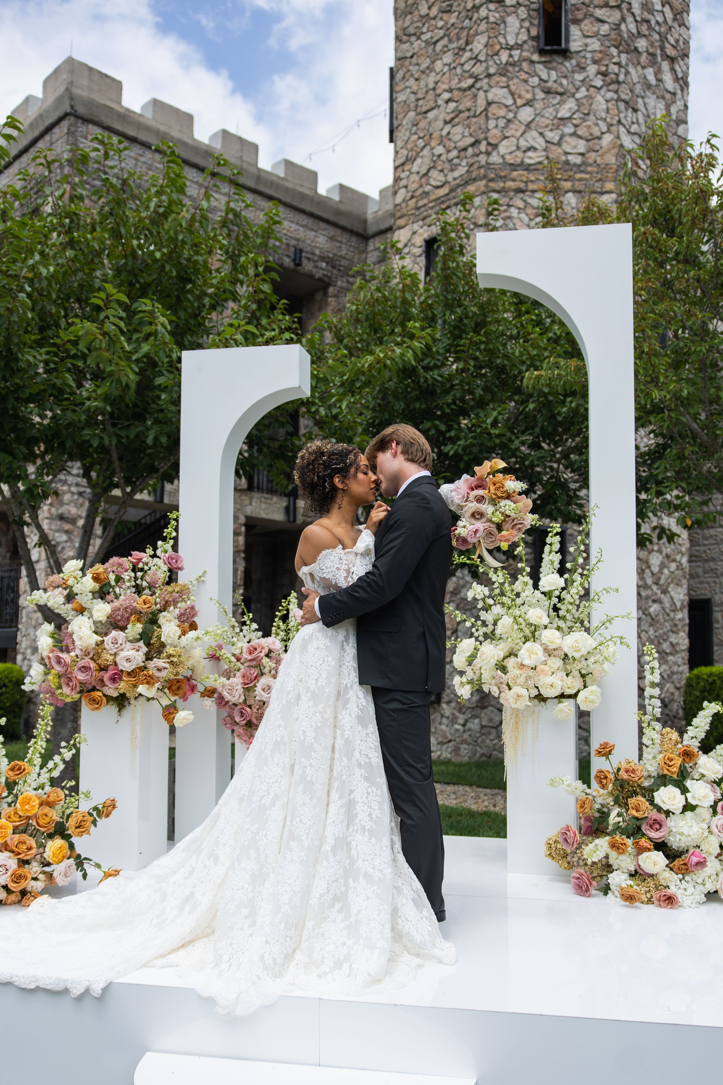Bride and groom share a romantic kiss at an outdoor wedding ceremony with floral arrangements and stone castle backdrop.