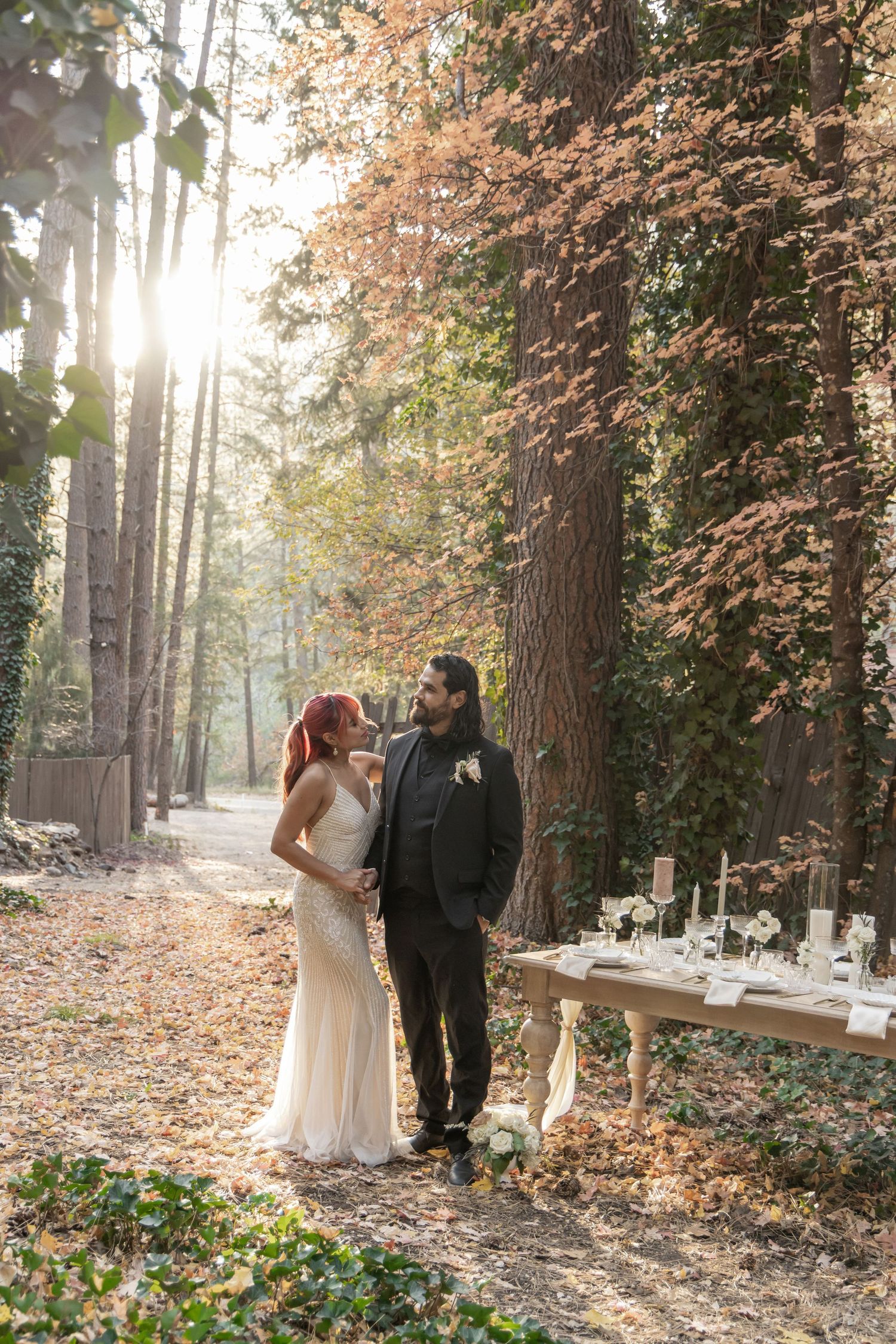 Bride in white gown and groom in black suit stand together in sunlit forest near elegant outdoor reception table.