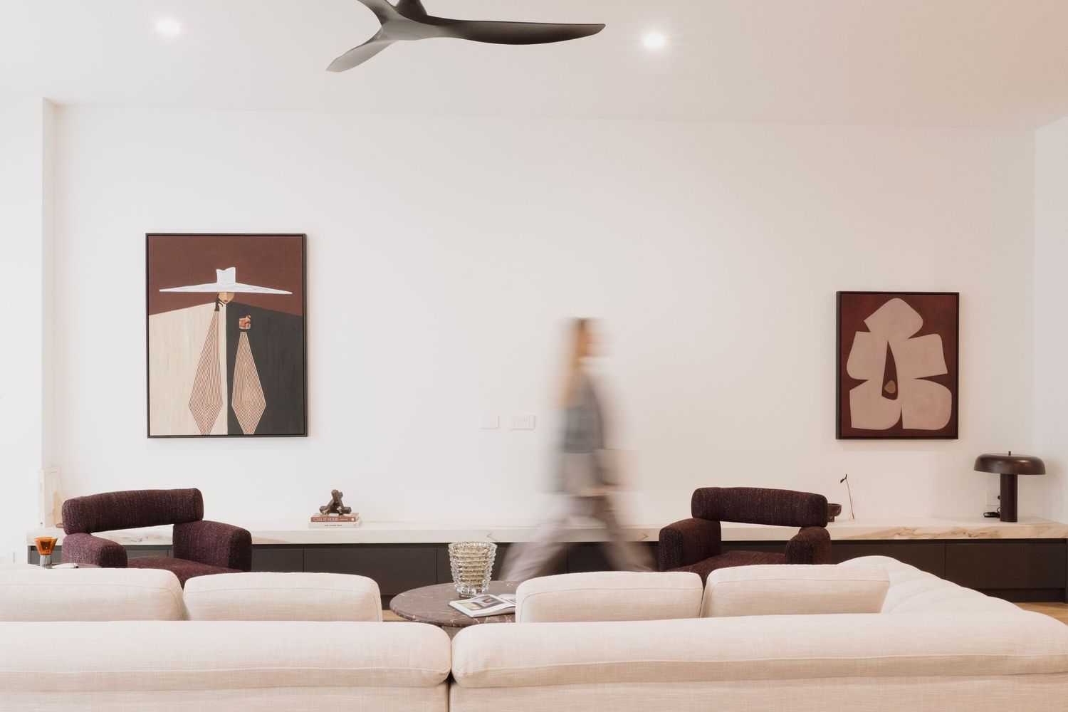Modern living room with white sofas, dark artwork, ceiling fan, and a blurred figure walking past.