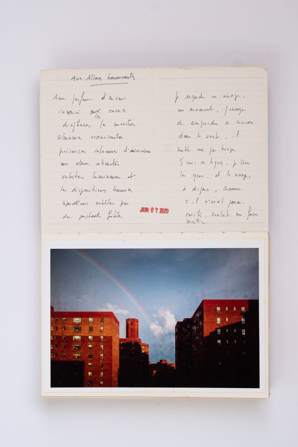 Open notebook with handwritten French text beside a photo of a rainbow over red brick New York City apartment buildings.