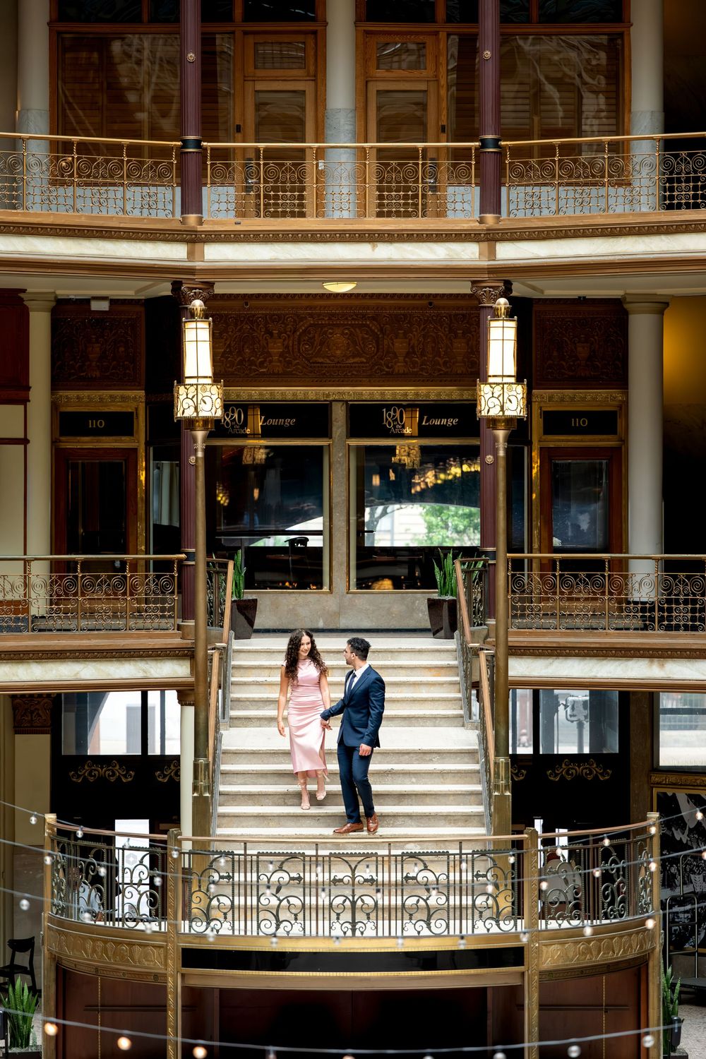 Elegant couple descends grand staircase in ornate historic building with gold balconies.