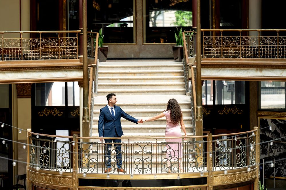 Couple holds hands on ornate balcony with grand staircase in elegant gold venue.