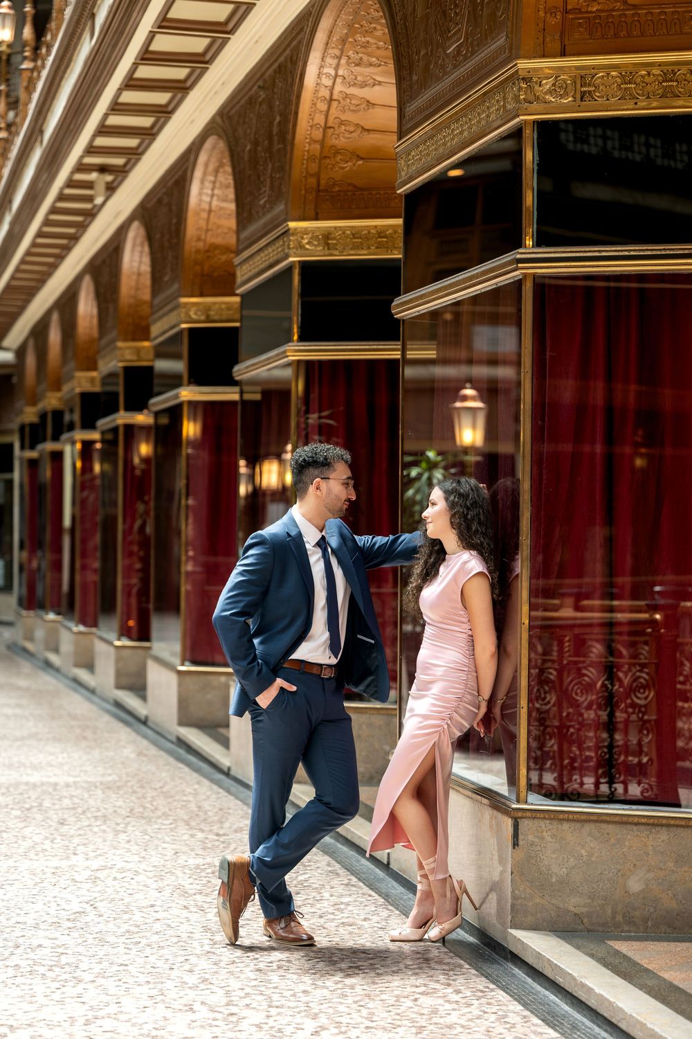 Stylish couple in formal attire sharing an intimate moment in an opulent gold and red corridor.