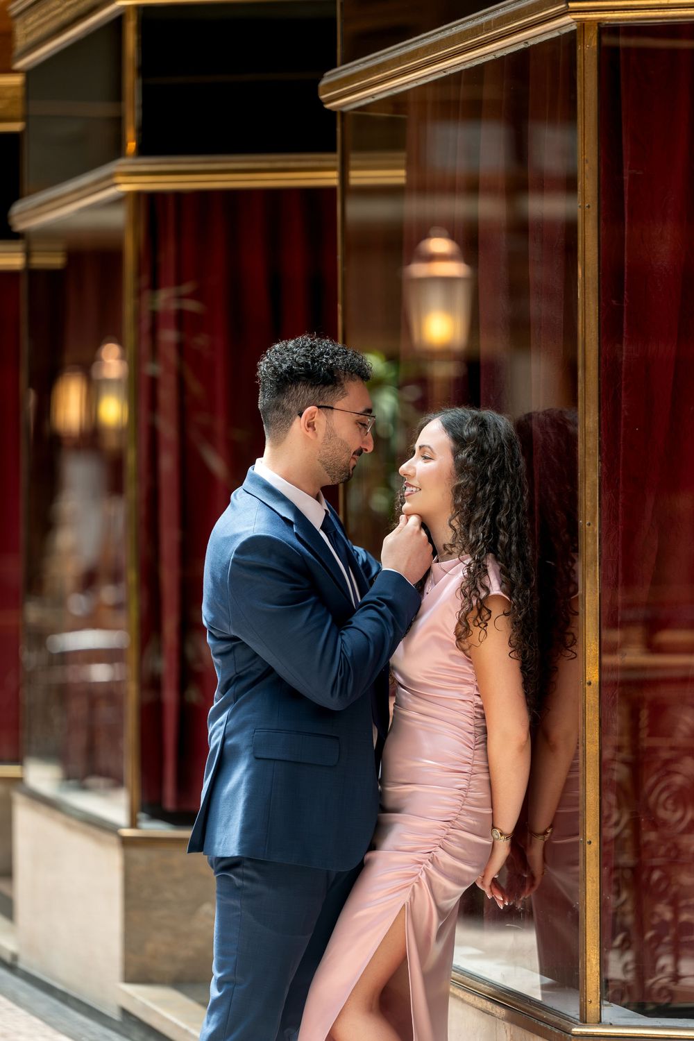 Couple in elegant attire share a romantic moment outside a restaurant, man in blue suit touches woman's face.