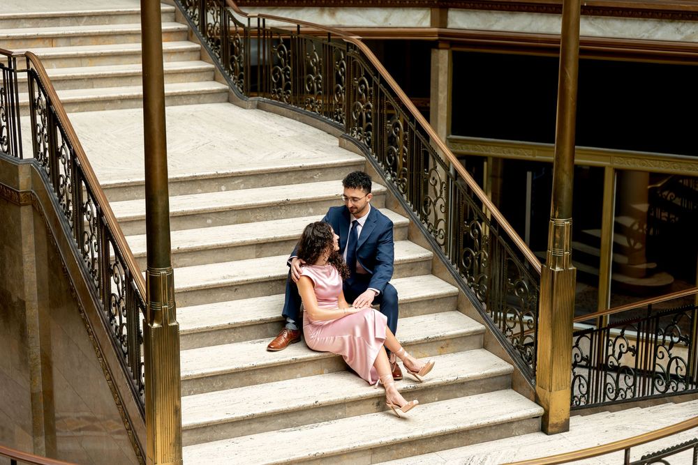 Couple sitting together on elegant marble staircase with ornate iron railings, woman in pink dress, man in navy suit.