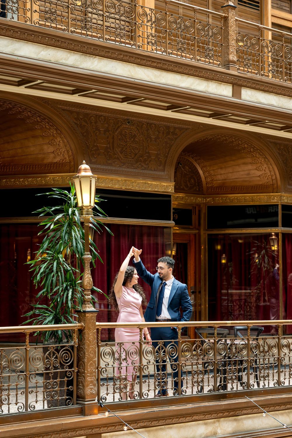 Couple dancing on ornate golden balcony of historic building with elegant arches and glowing lamp post.