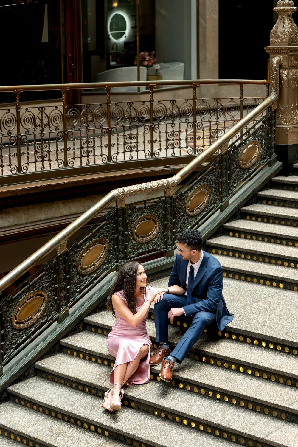 Couple sits together on grand ornate staircase with decorative gold railings in historic building.