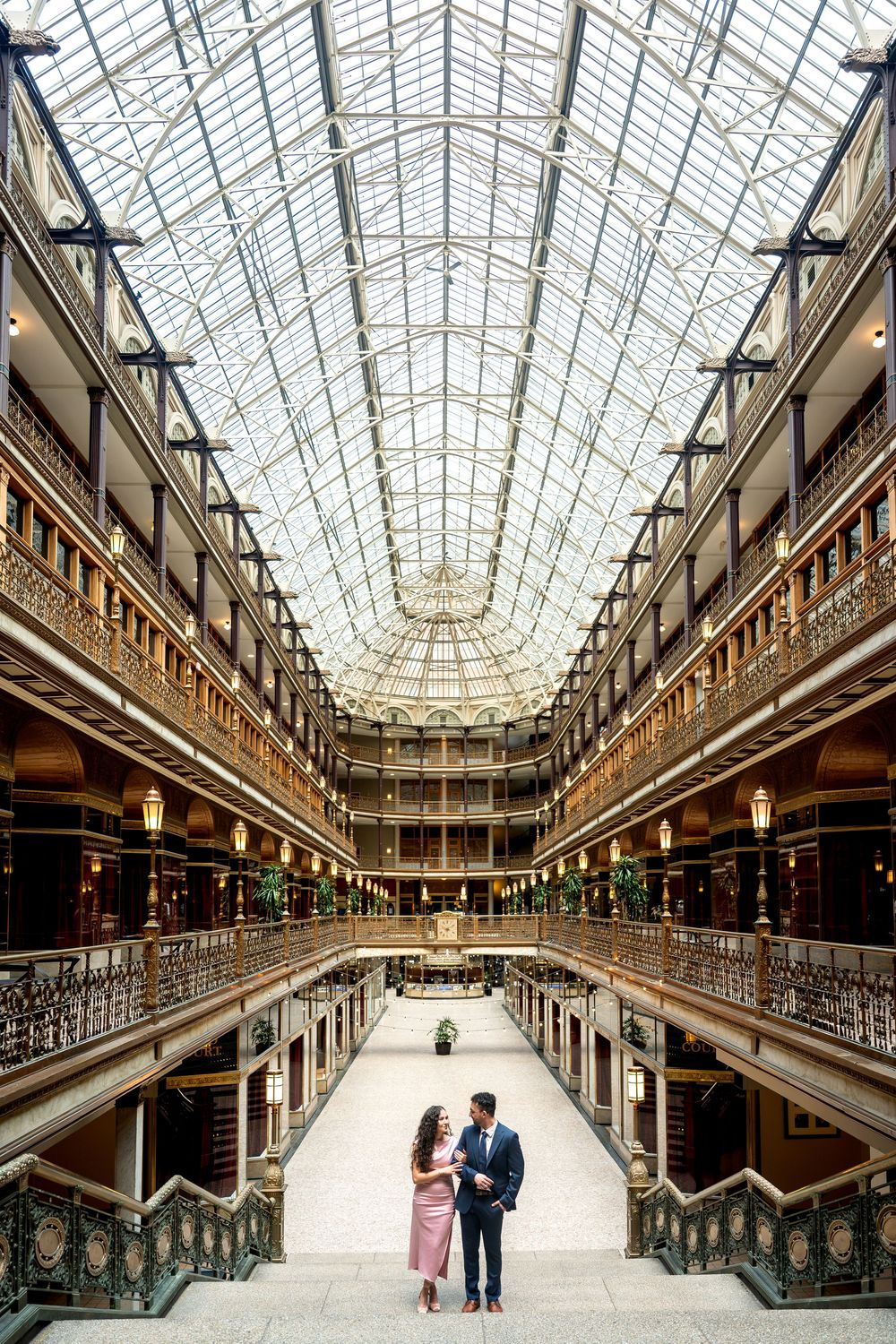 Couple stands together in stunning historic arcade with soaring glass skylight ceiling above.
