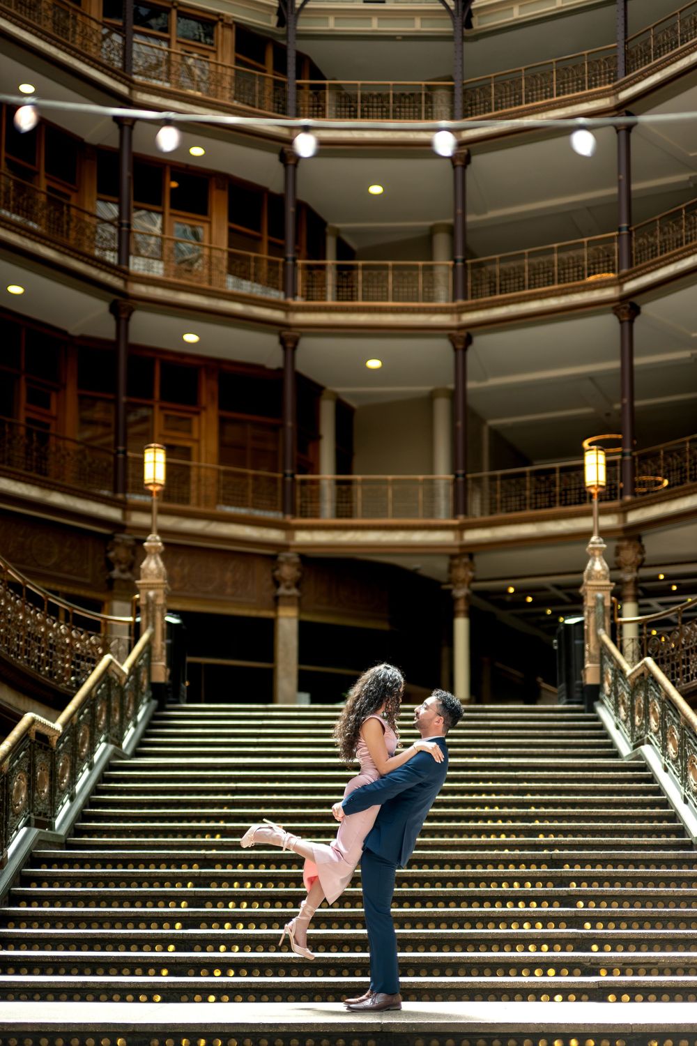 Man lifts woman on grand staircase inside stunning multi-tiered historic building with warm lights.