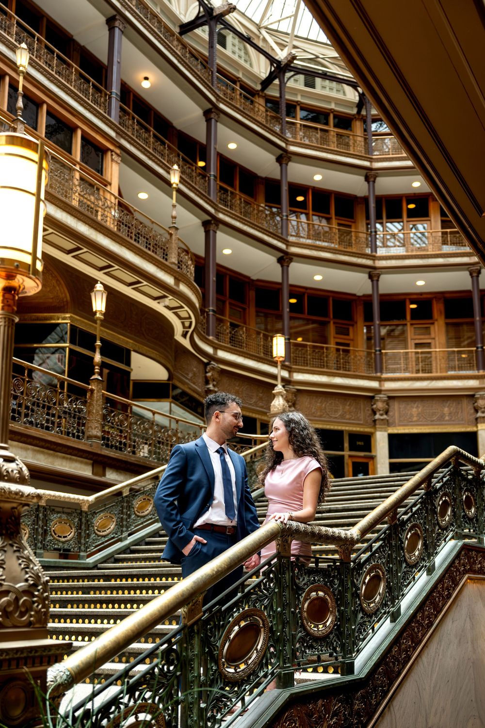 Elegant couple in formal wear share a romantic moment on ornate stairs inside a grand historic building.