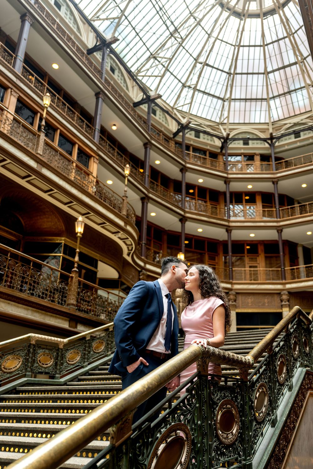 Couple sharing a kiss on ornate staircase inside a stunning historic building with glass dome ceiling.