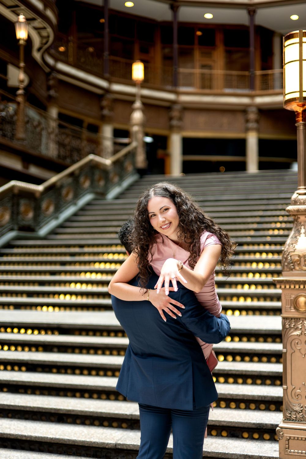 Smiling woman shows engagement ring while being lifted by partner on ornate historic staircase.