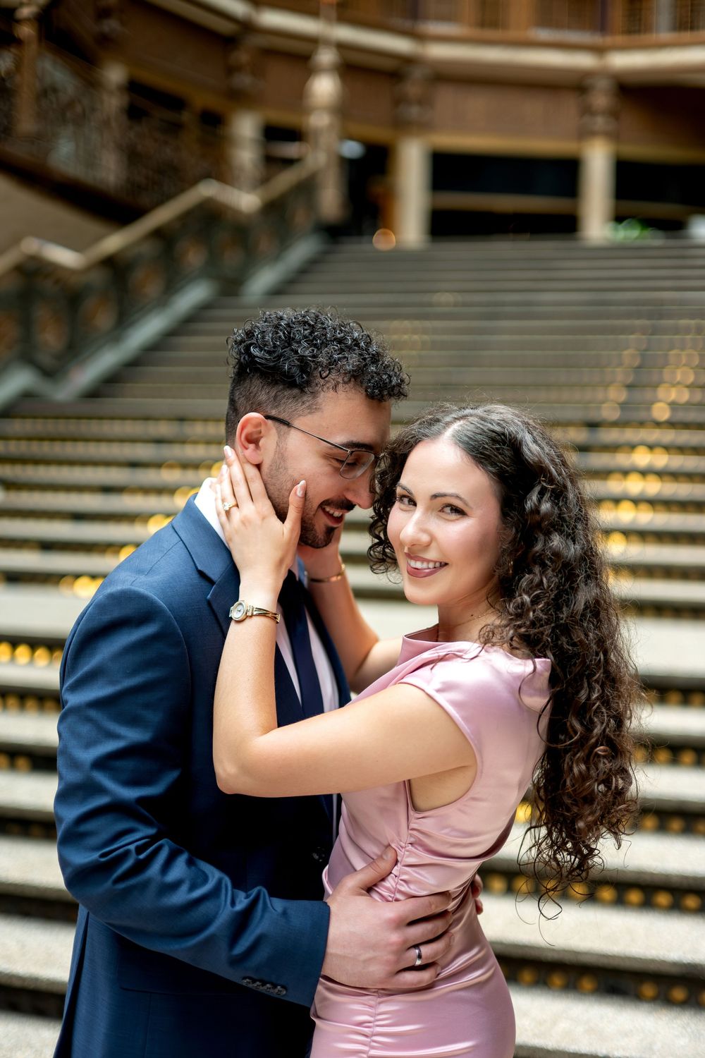 Smiling woman in pink satin dress embracing man in blue suit on grand staircase indoors.