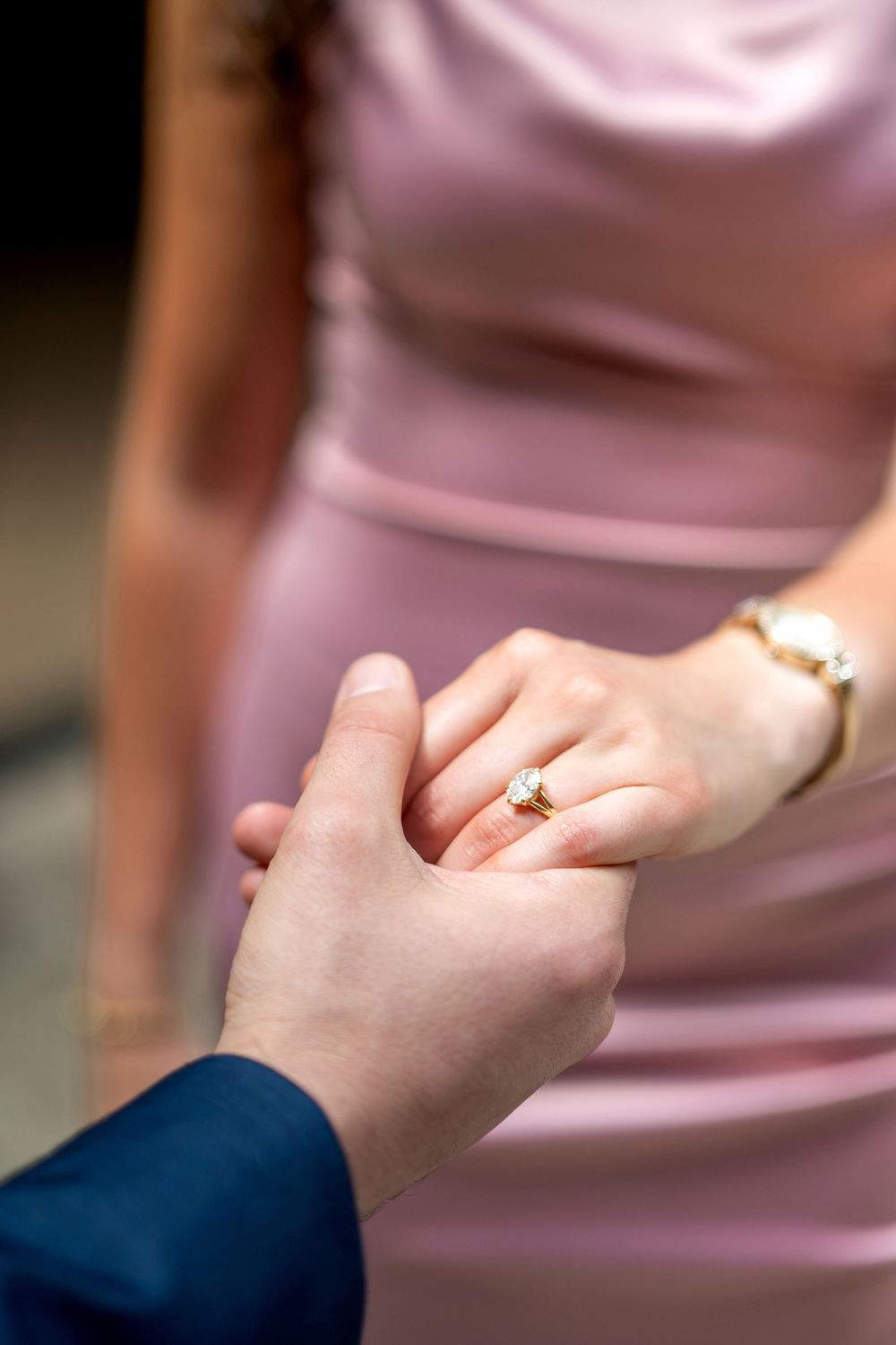 Close-up of woman's hand wearing a diamond engagement ring held by man in navy suit sleeve.