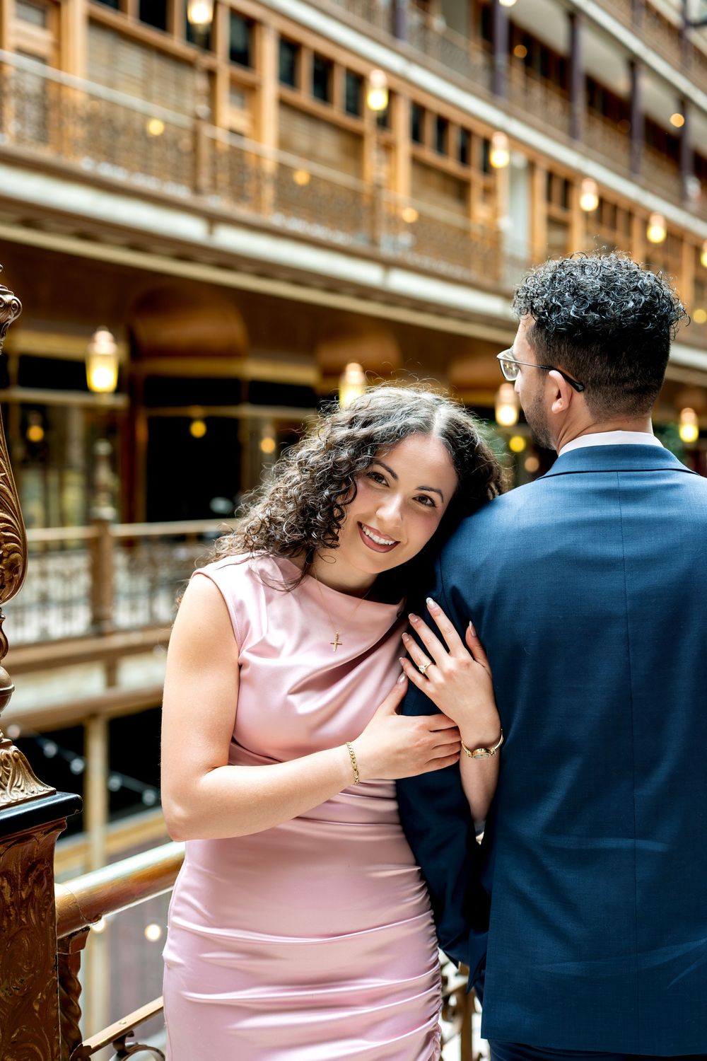 A smiling woman in a pink satin dress embraces a man in a navy suit on a balcony of an elegant building.