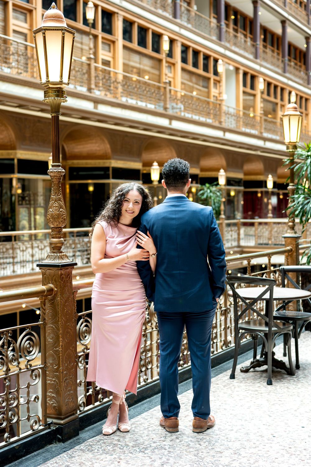 Couple in elegant attire poses by ornate gold railing in a grand historic building interior.