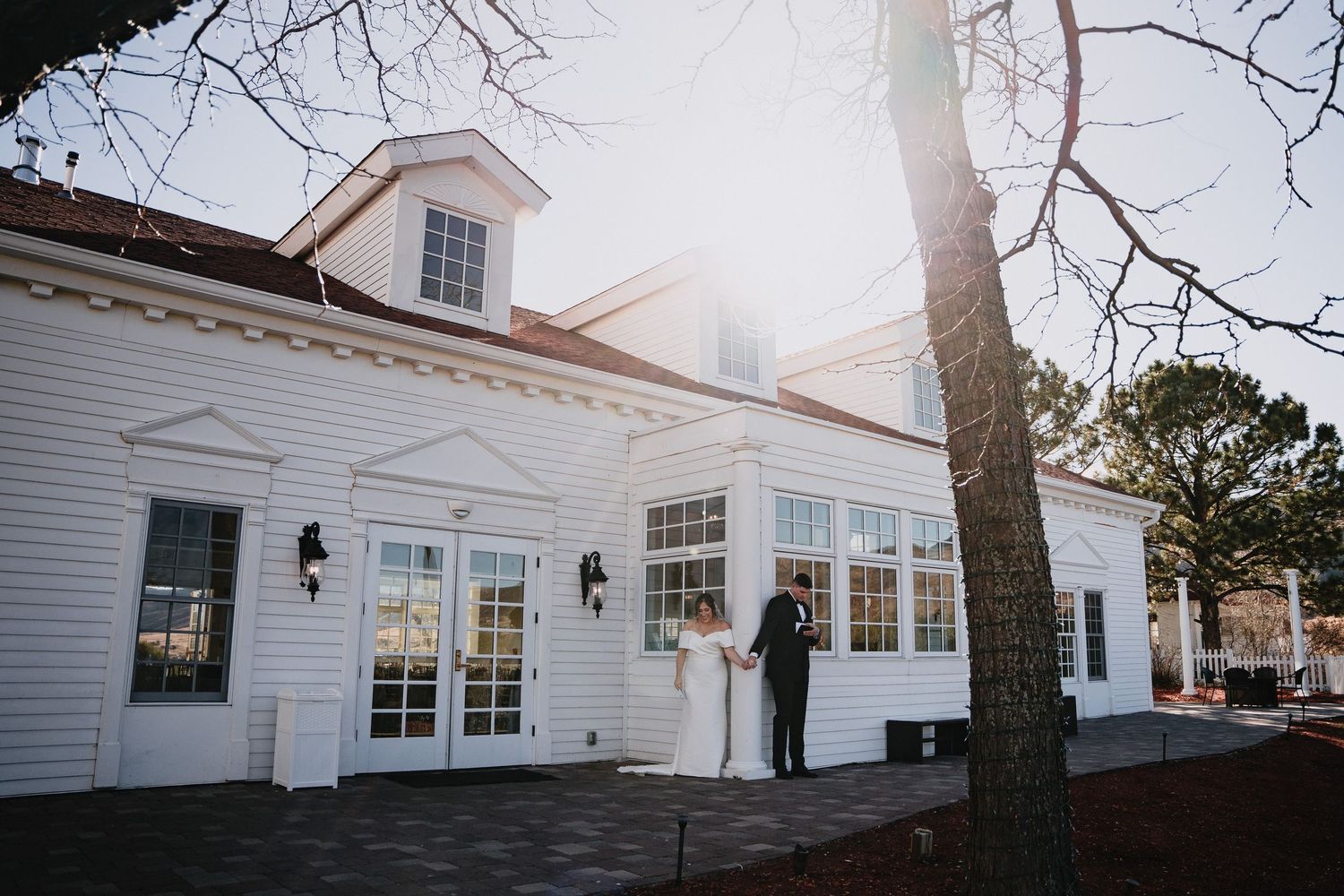 Bride and groom stand outside a charming white colonial wedding venue with large windows and a sunlit tree.