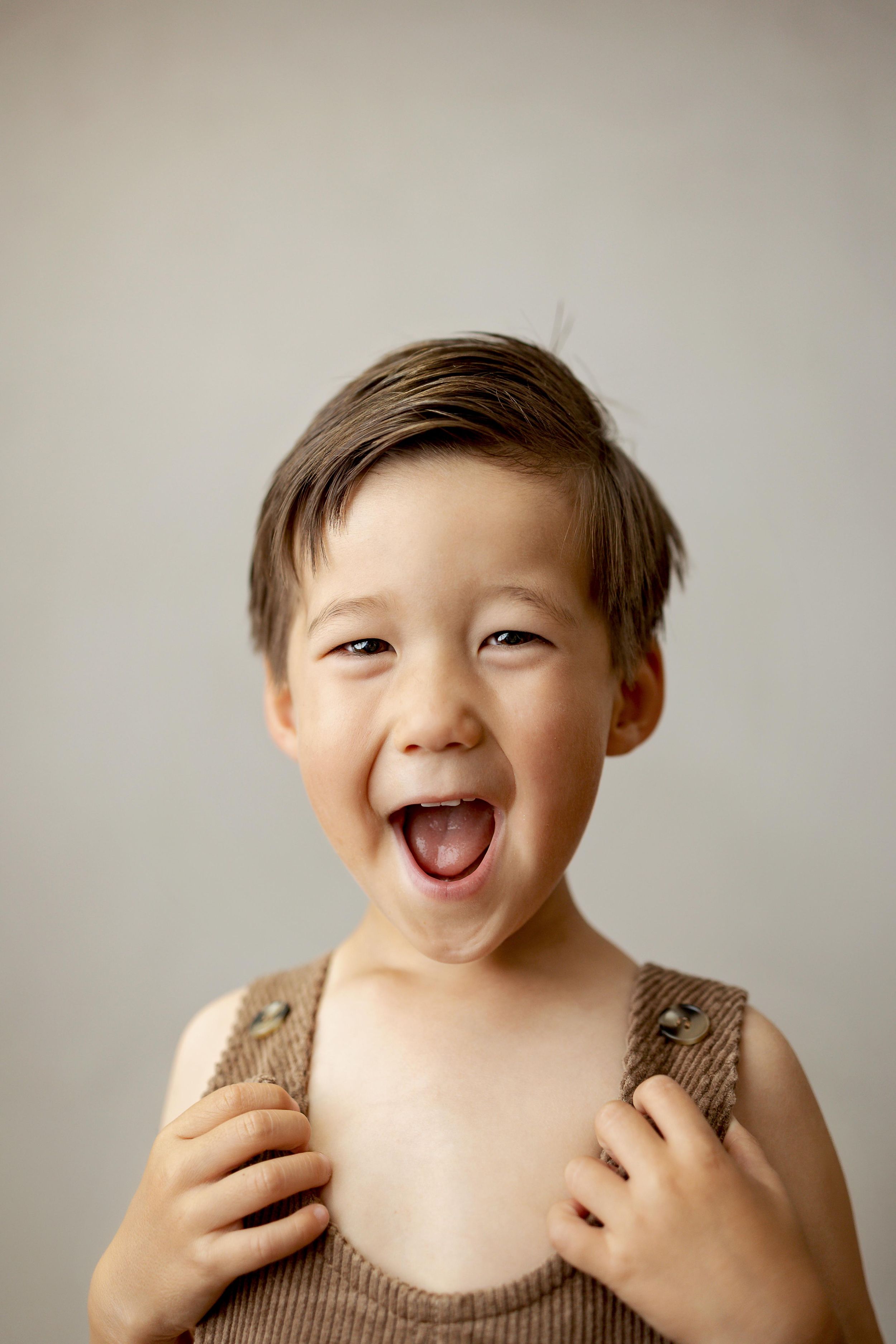Happy young boy laughing with mouth open, wearing a brown knit tank top, portrait against neutral background.