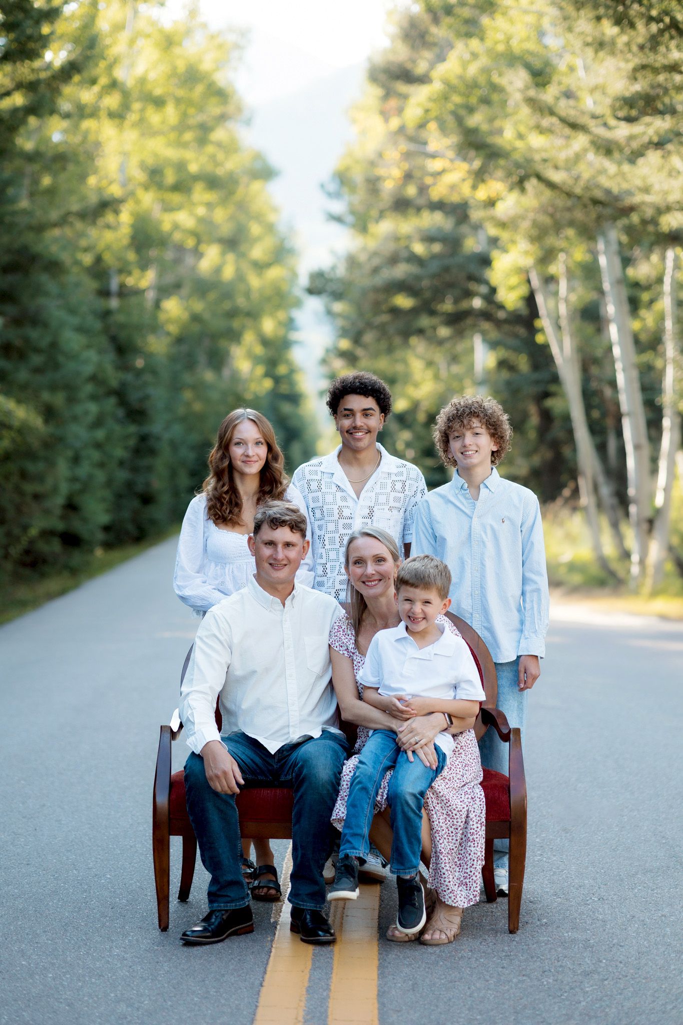 Family of six posing on a tree-lined road, seated and standing around a wooden bench in casual summer outfits.