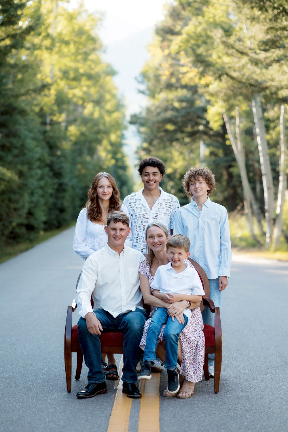 Family of six posing on a tree-lined road, seated and standing around a wooden bench in casual summer outfits.