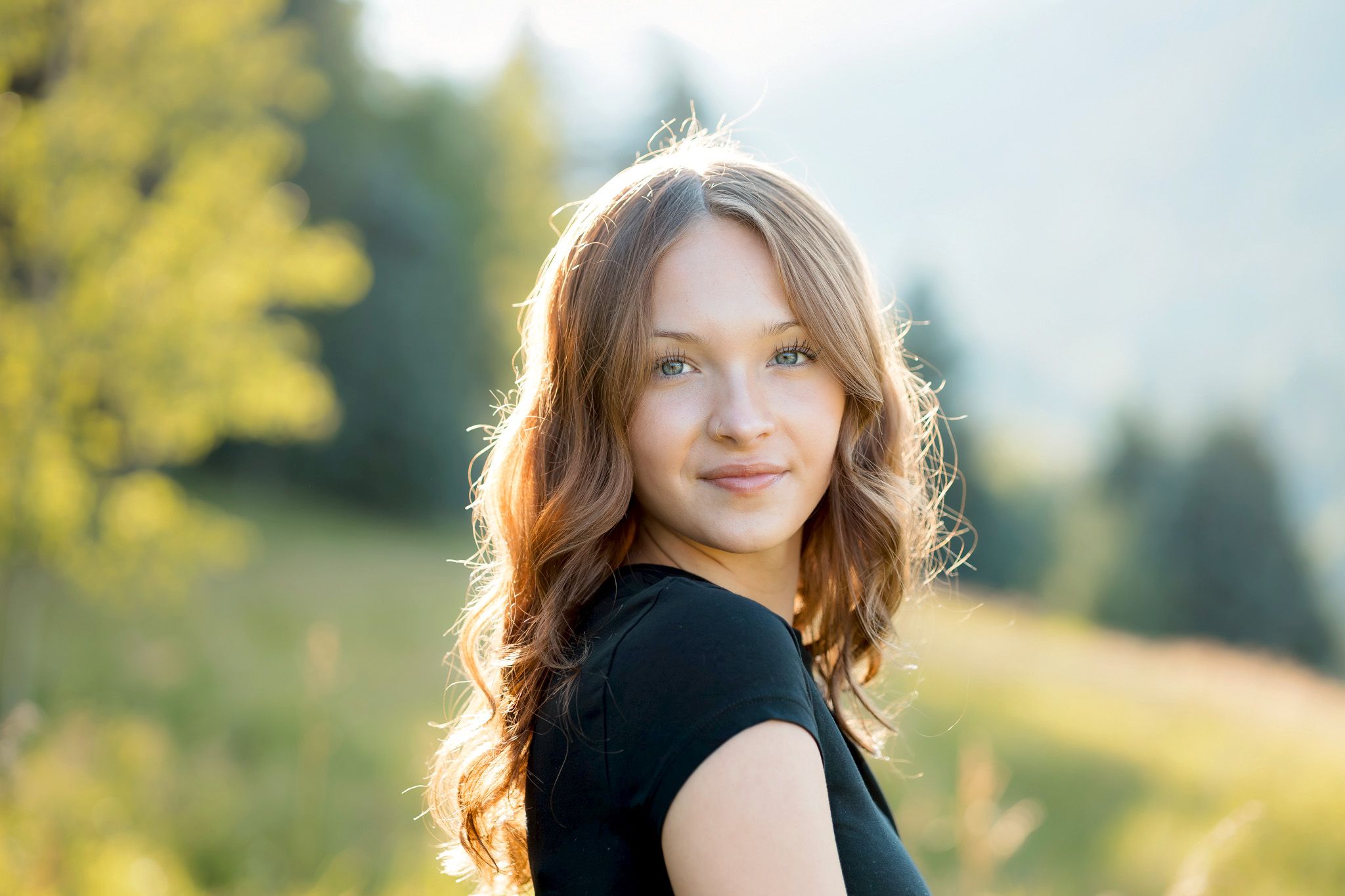 Young woman with long auburn hair smiling outdoors in golden sunlight with green hills in the background.