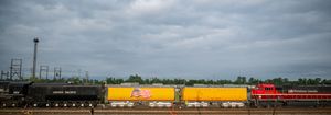 A vintage steam locomotive alongside yellow freight train cars on railroad tracks under a cloudy sky.