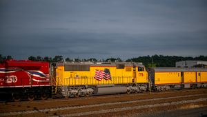 Yellow Union Pacific locomotive with American flag livery leads a freight train under cloudy skies at a rail yard.