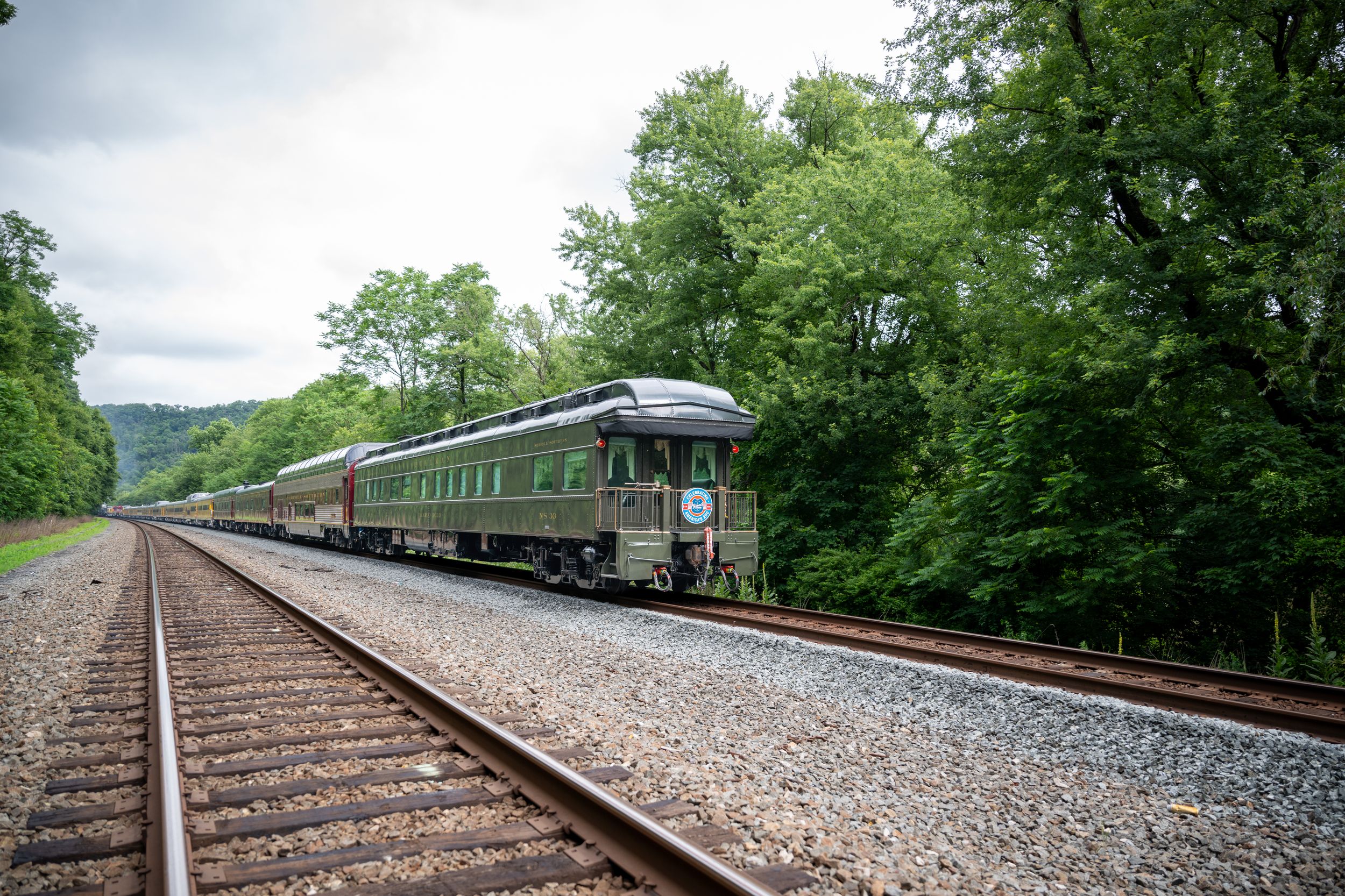 Vintage passenger train traveling through lush green forest on a scenic railway track on a cloudy day.
