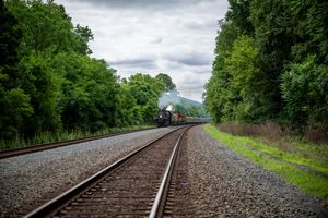 A vintage steam train travels along railroad tracks surrounded by lush green trees under a cloudy sky.