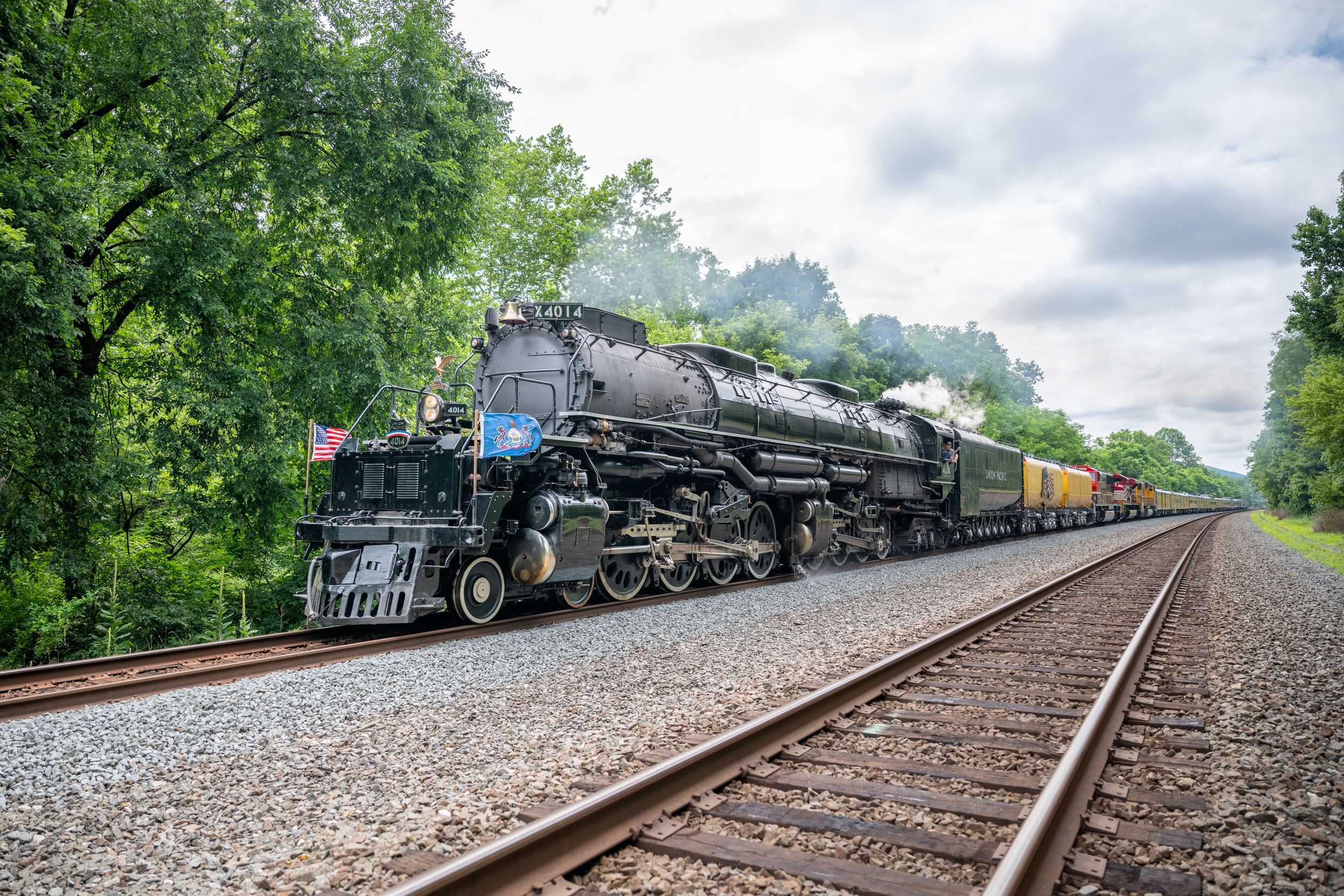 A vintage black steam locomotive pulls freight cars along a tree-lined railway under a cloudy sky.