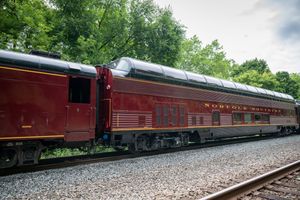 Vintage Norfolk Southern red passenger train cars on railroad tracks surrounded by lush green trees.