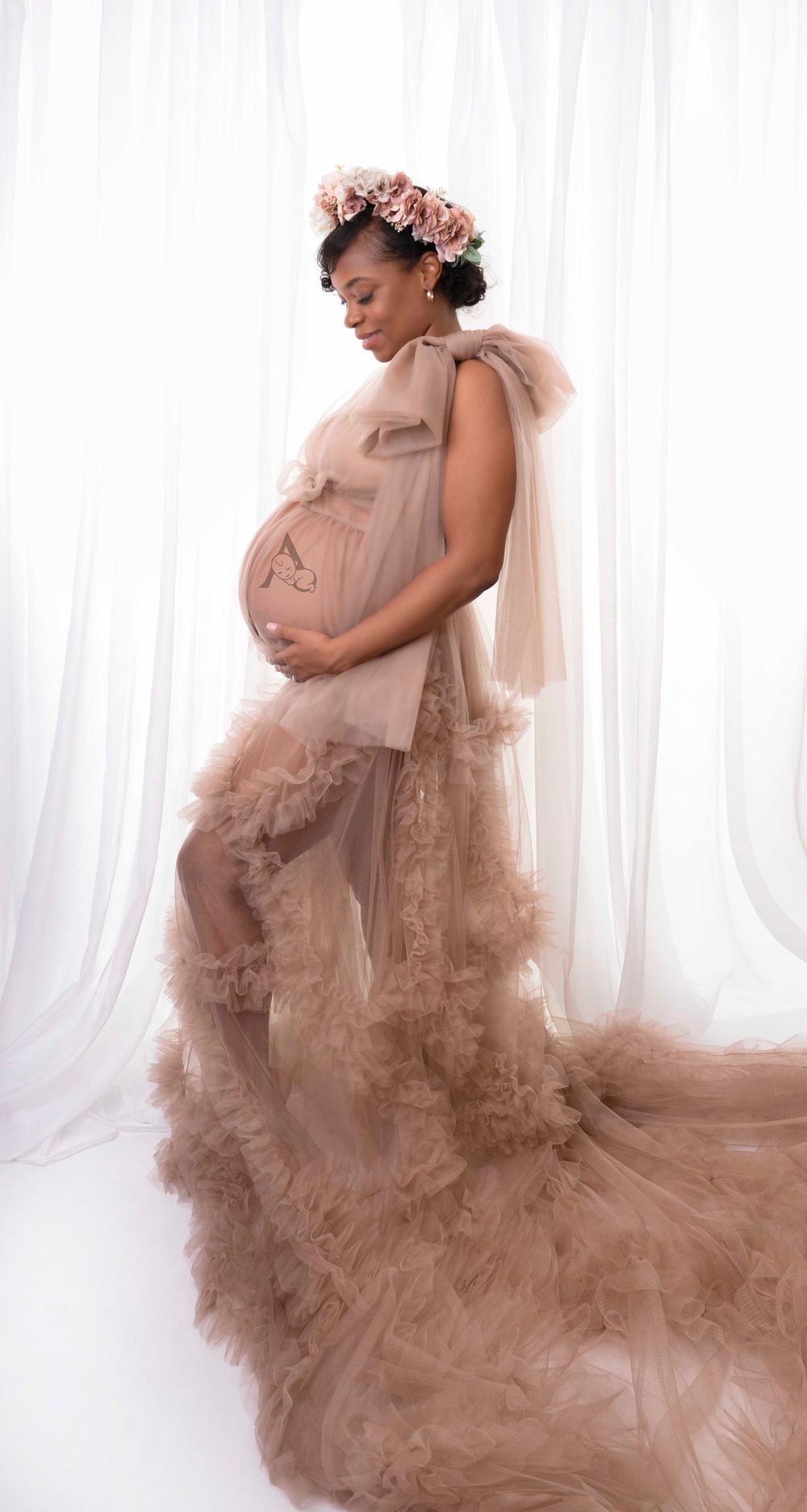 Maternity portrait in flowing blush gown with floral crown against white backdrop.
