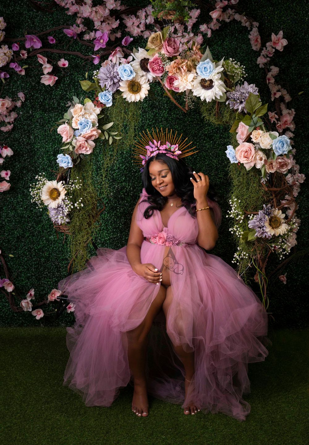 A dreamy maternity photoshoot with a pink tulle gown under a floral arch against a dark green backdrop.