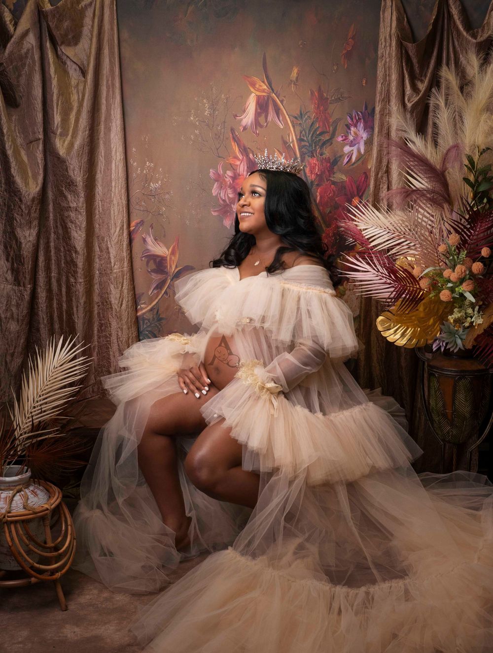 Elegant maternity portrait with cream tulle gown and floral decor against draped fabric backdrop.