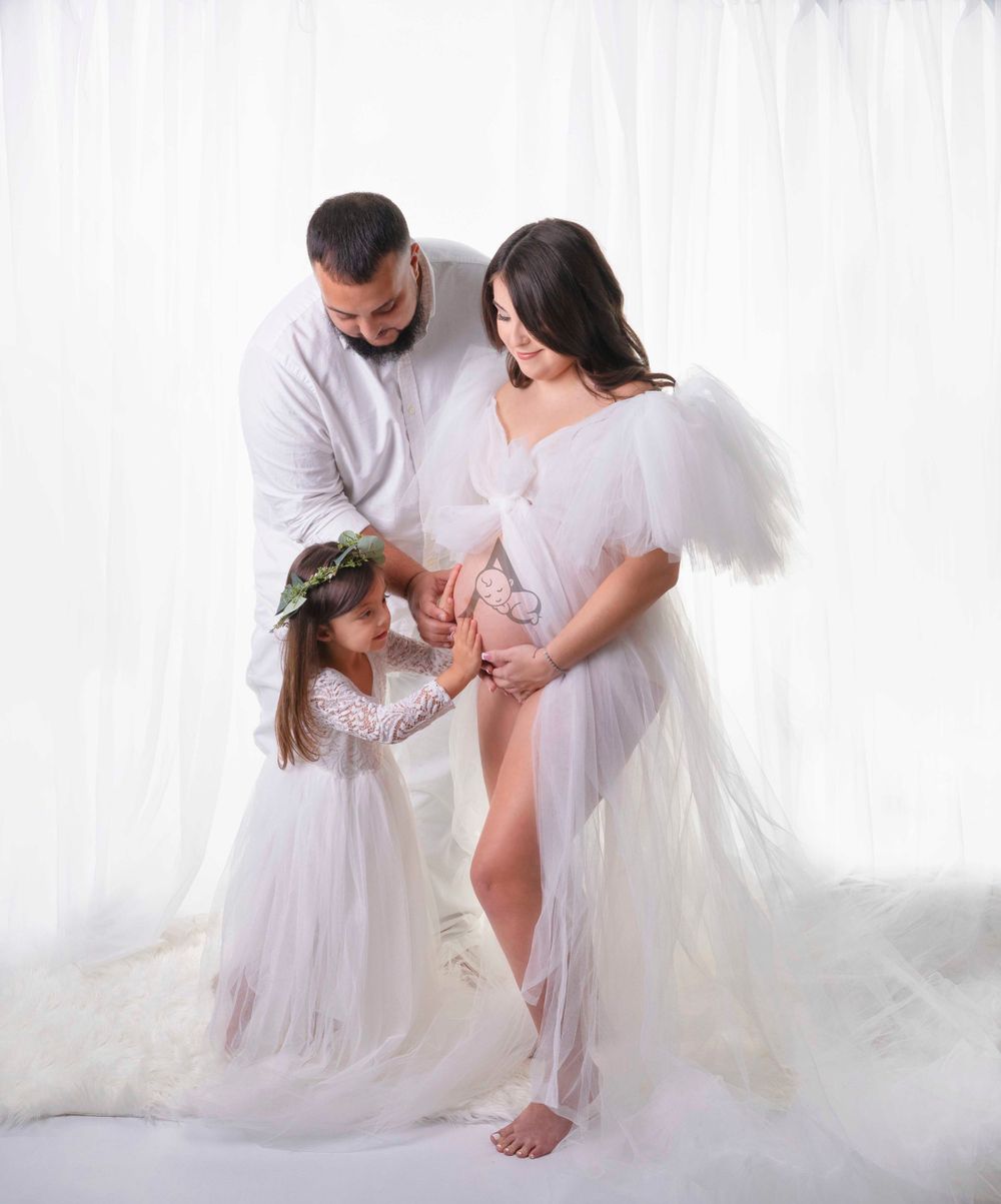 Ethereal family maternity portrait with white wings and flowing fabrics against white backdrop.