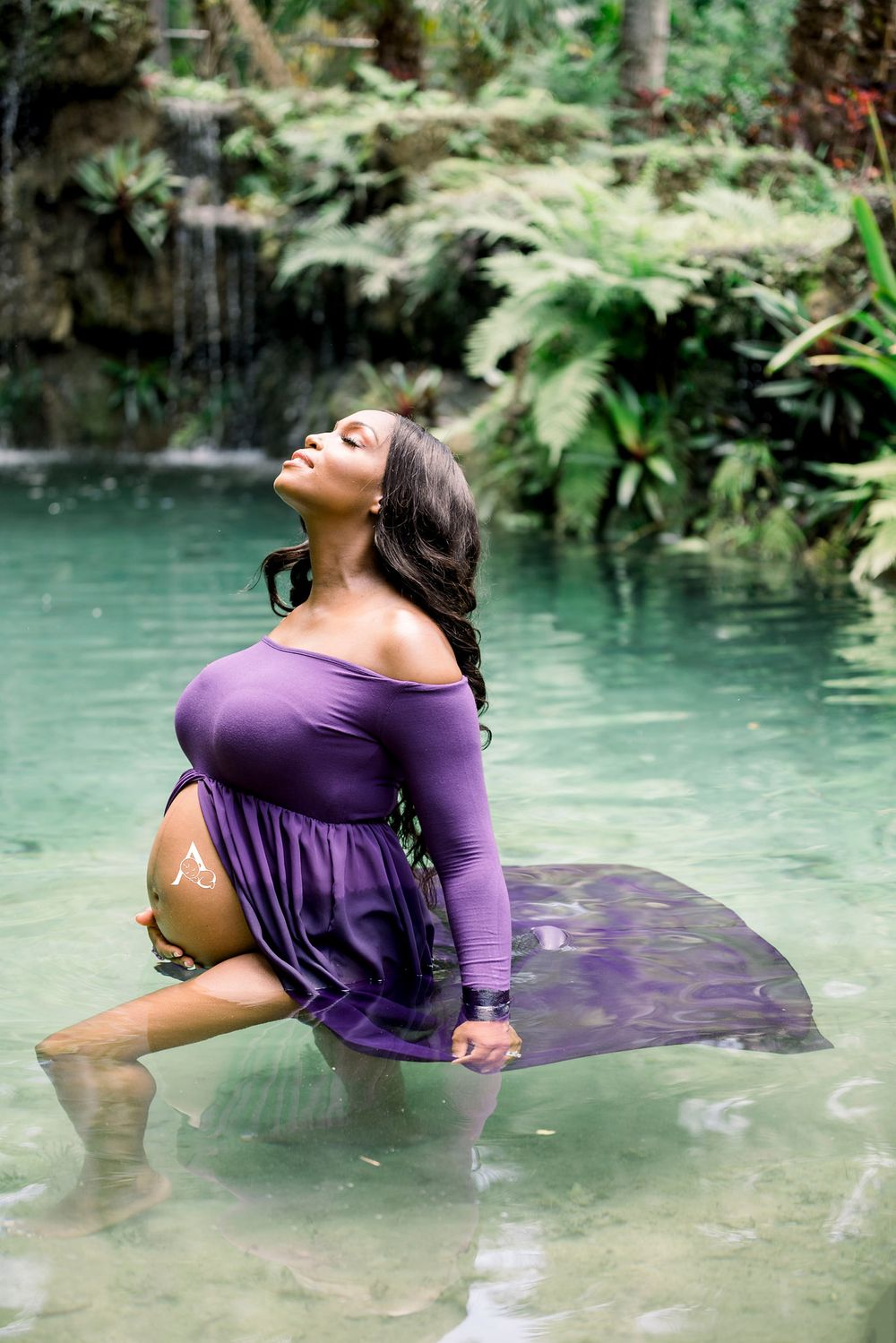 Artistic maternity photo in purple dress with subject posed in shallow turquoise waters.