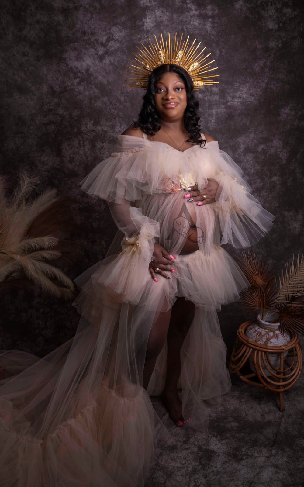 Ethereal maternity portrait featuring a woman in white tulle gown with golden crown against dark backdrop.