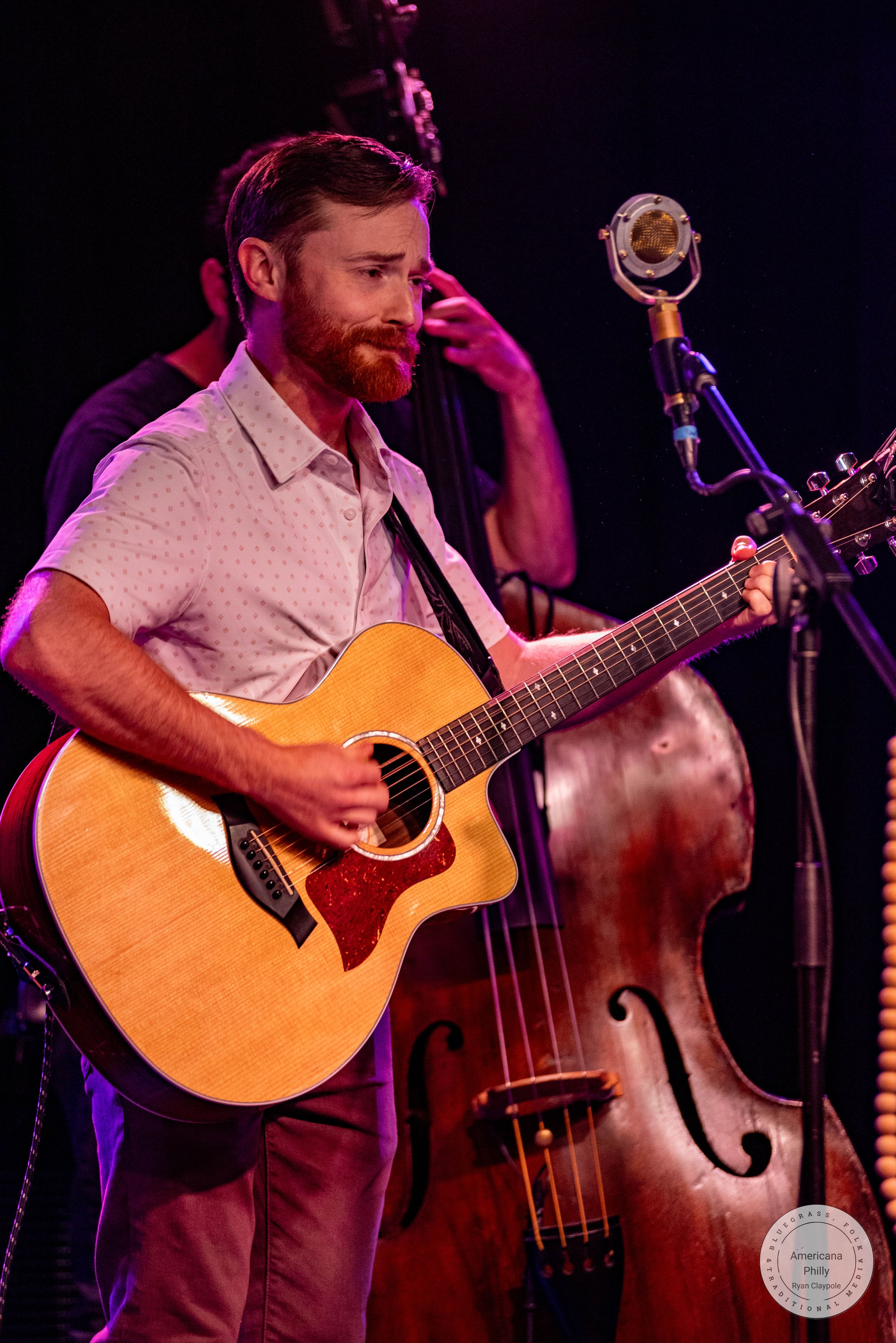 Bearded man playing acoustic guitar on stage near a vintage microphone with a double bass player in the background.