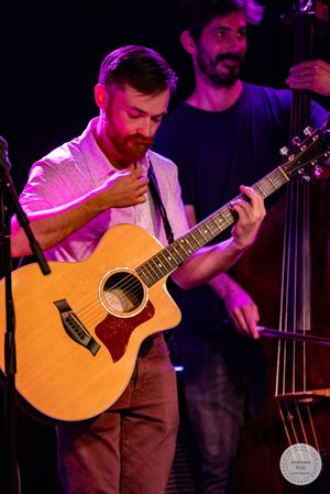 Musician playing acoustic Taylor guitar on stage with upright bass player in background under purple stage lighting.