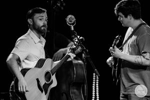Two musicians perform on stage in black and white, one playing acoustic guitar and another electric guitar near a vintage microphone.