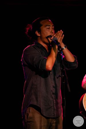 A young Asian male singer performs on stage, holding a microphone with both hands under dramatic red and purple stage lighting.