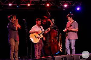 Four musicians perform on stage with guitar, upright bass, trumpet, and banjo under colorful stage lights.