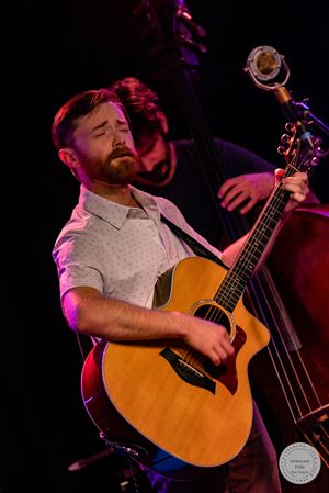 A bearded man plays acoustic guitar on stage with a double bass player visible behind him.