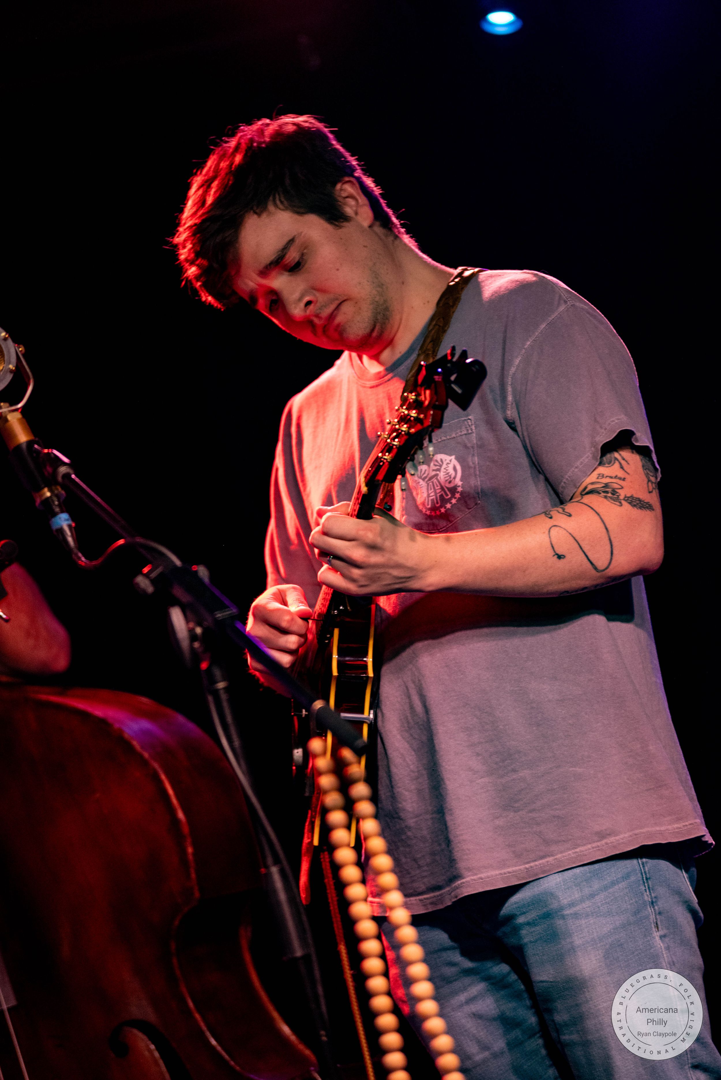 A tattooed young man plays electric guitar on a dark stage with a double bass nearby.