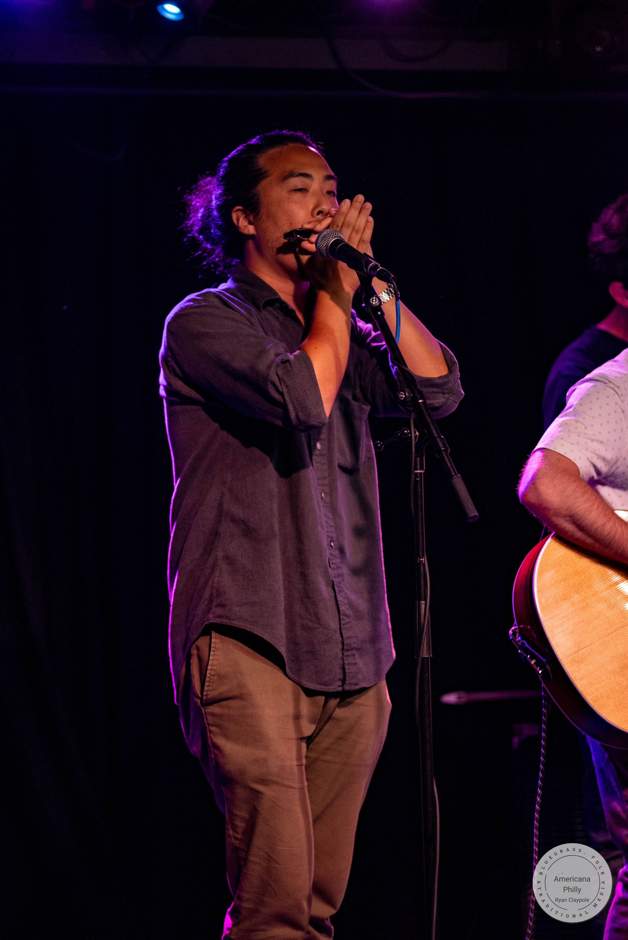 Male singer performing on stage at microphone, wearing gray shirt and khaki pants in dimly lit venue.