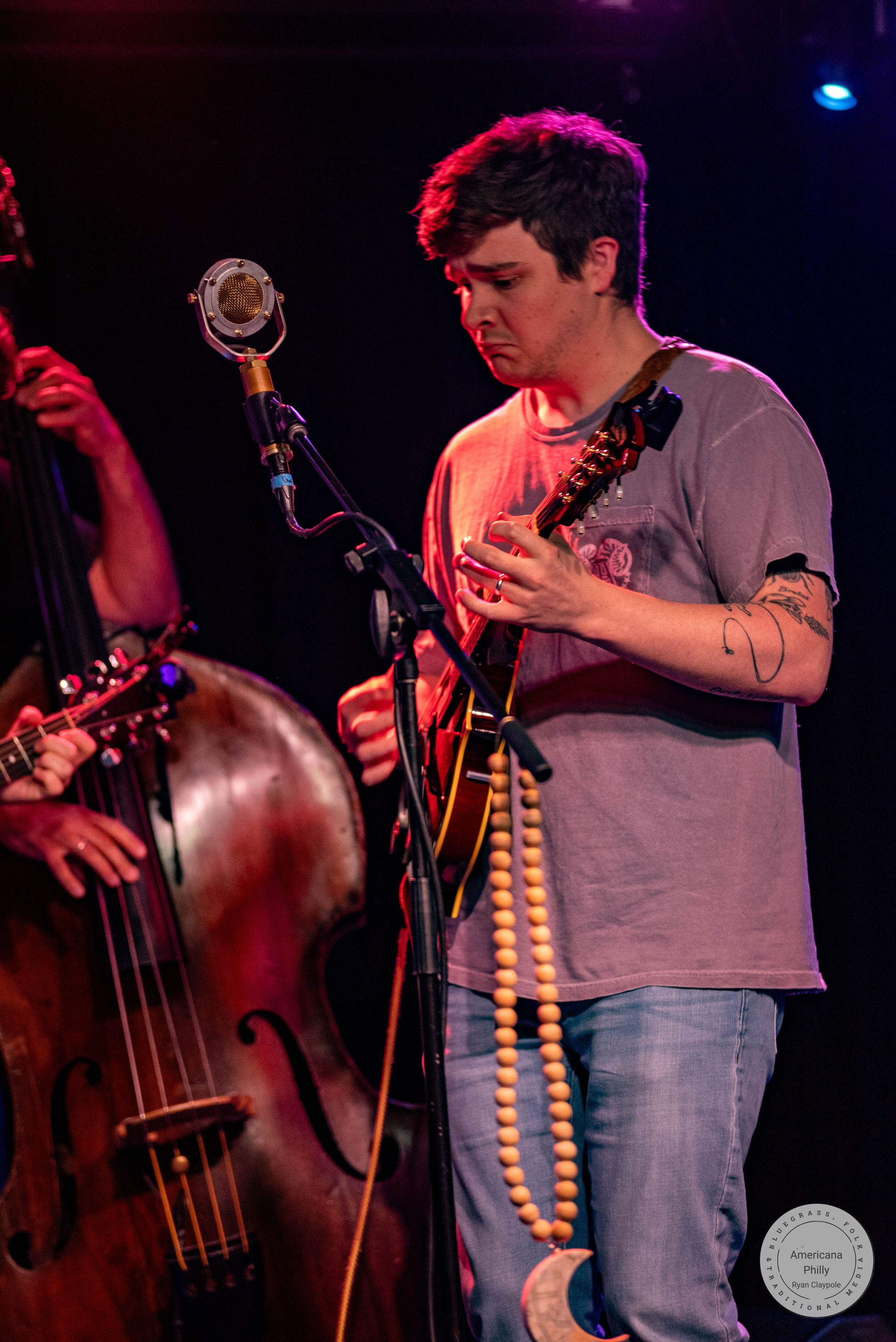 Musician playing mandolin on stage with a double bass player beside him under red stage lighting.