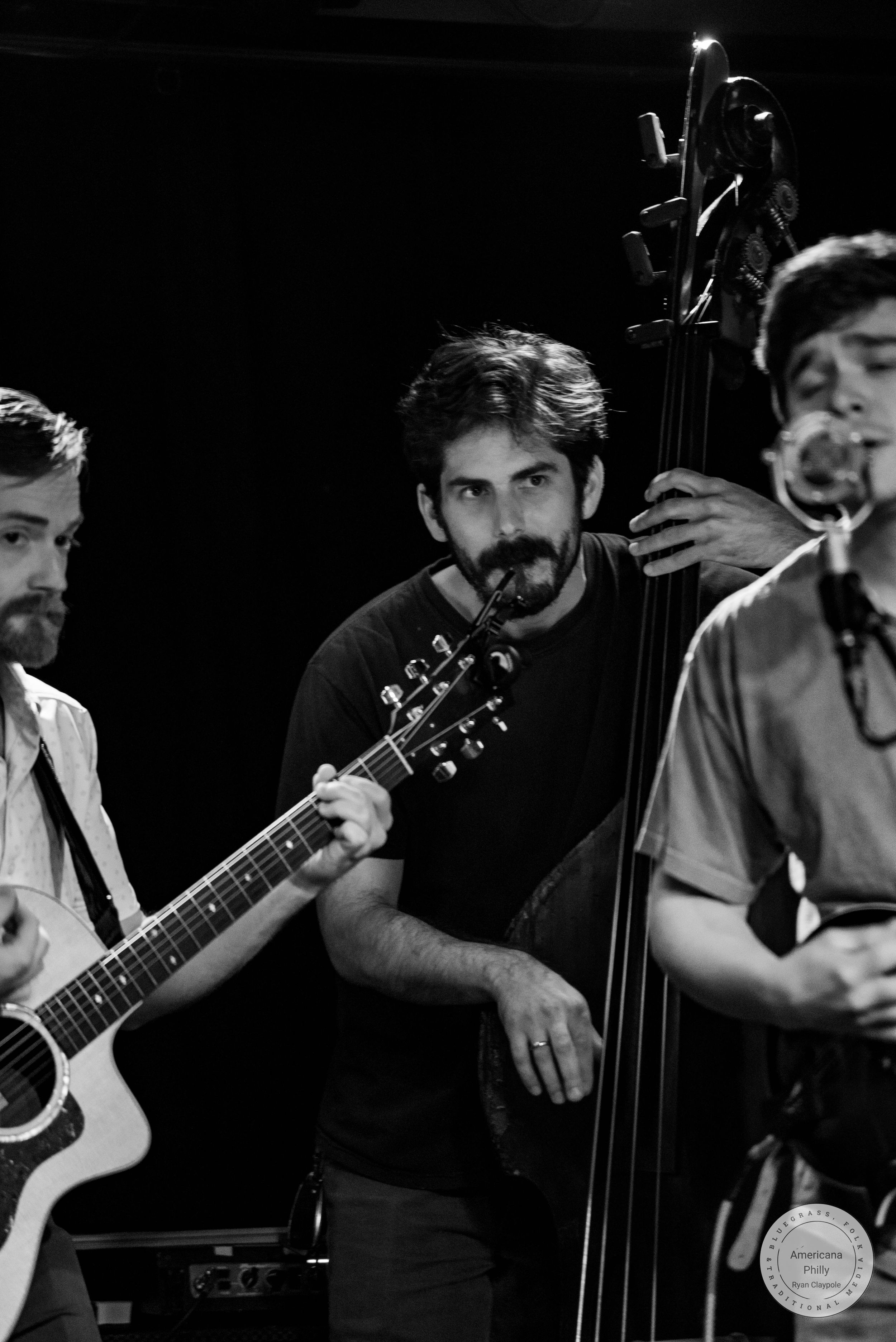 Black and white photo of three musicians performing live, featuring guitar and upright bass players on stage.