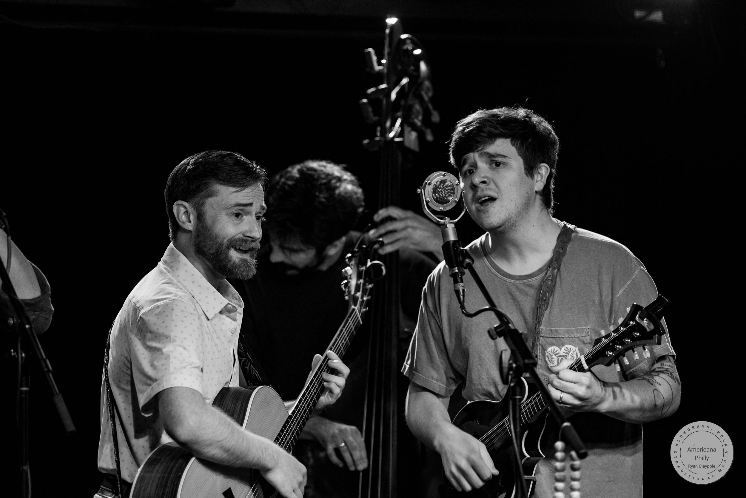 Two musicians smile and sing together on stage, playing acoustic guitars in a black and white live performance photo.