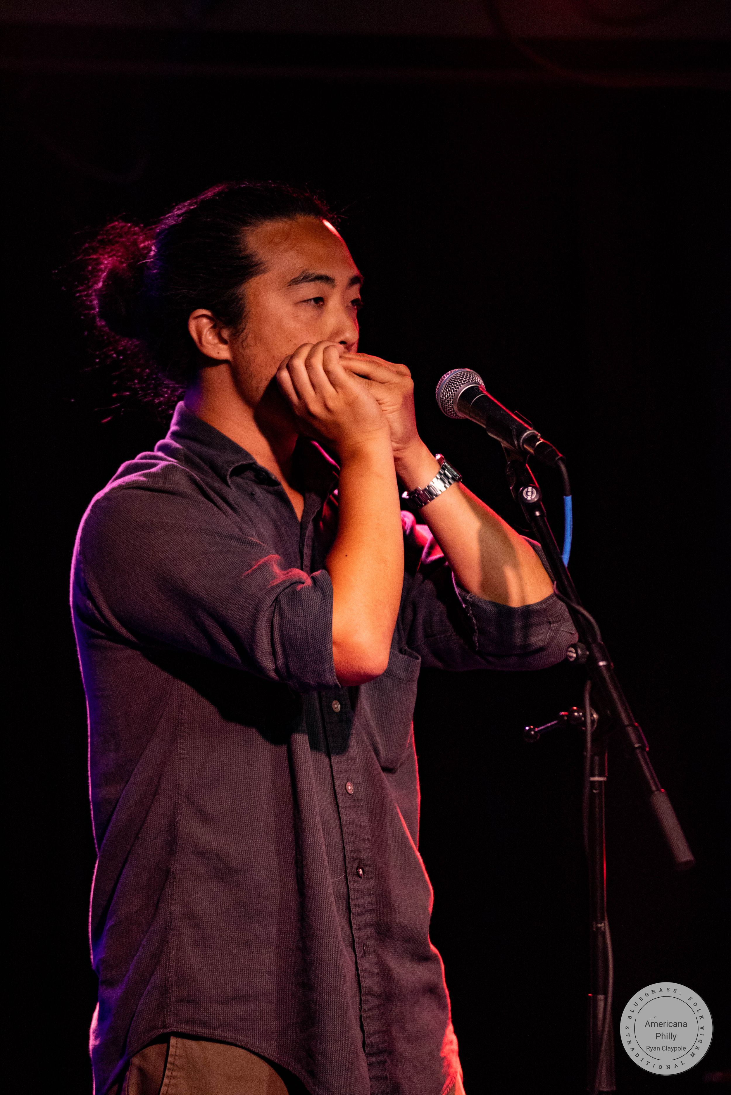 A young Asian man with a bun plays harmonica at a microphone stand on a dimly lit stage.
