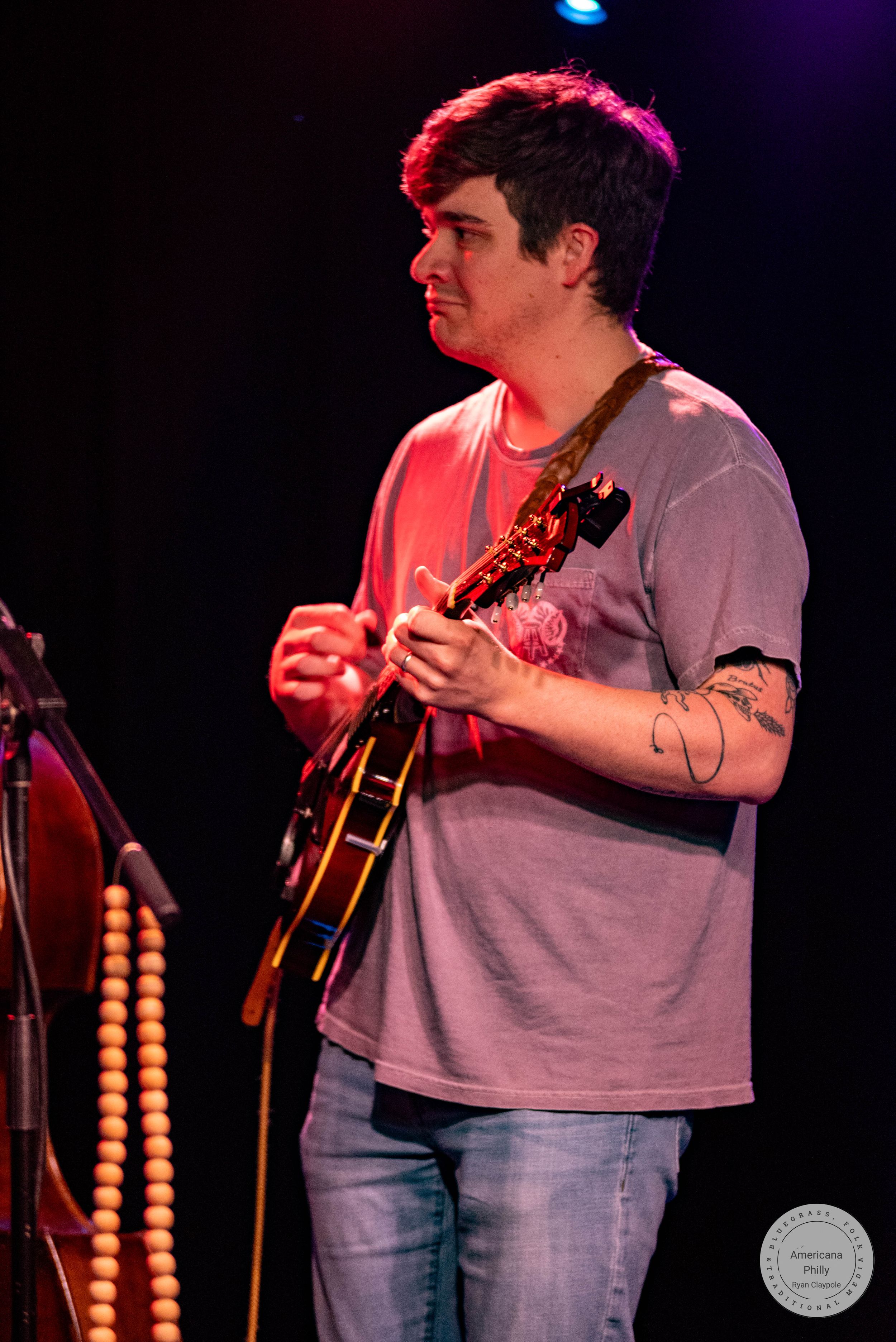 Young man playing electric guitar on stage under pink lighting, wearing gray t-shirt and jeans.